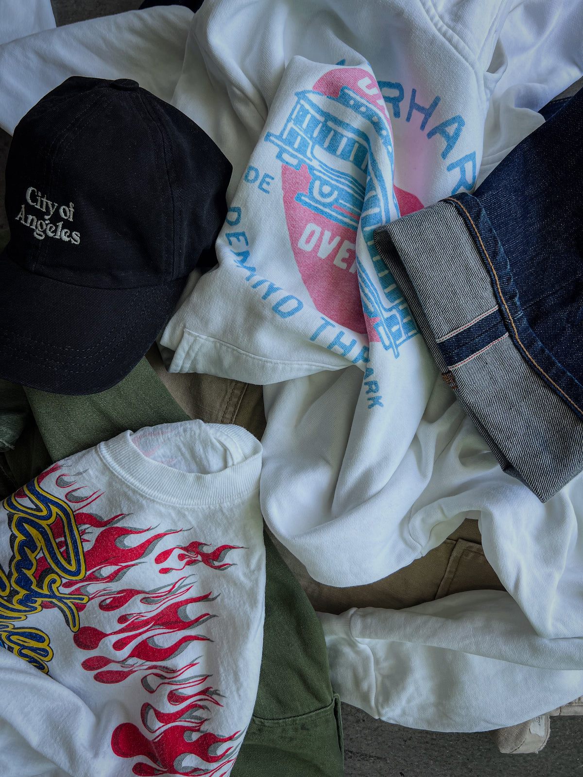 Vintage clothing flat lay with graphic T-shirts, selvedge denim, military pants, and a black cap.
