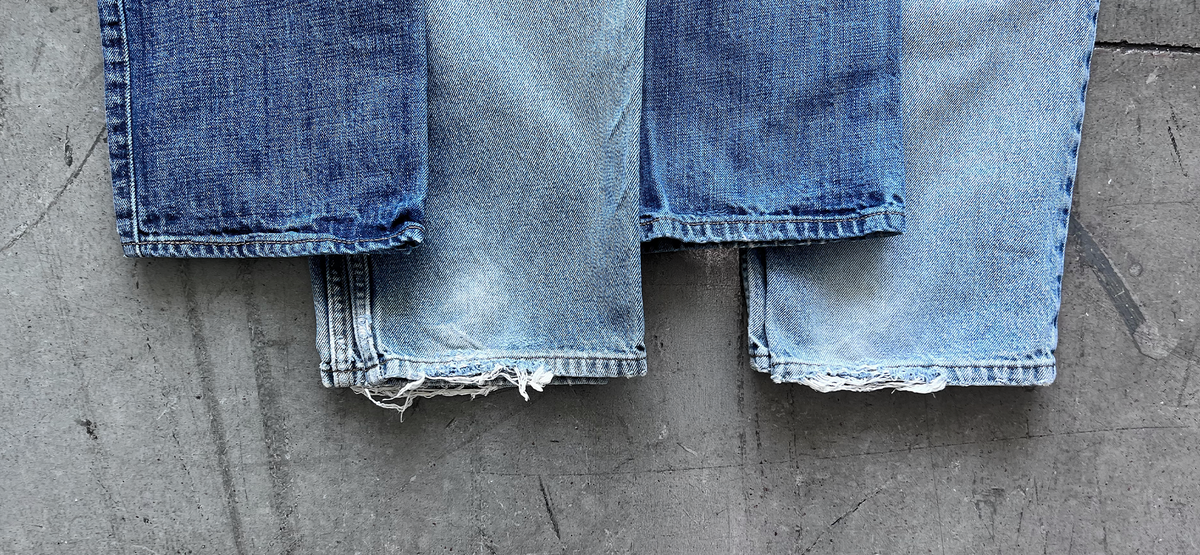Three pairs of worn blue jeans with frayed hems laid on concrete.