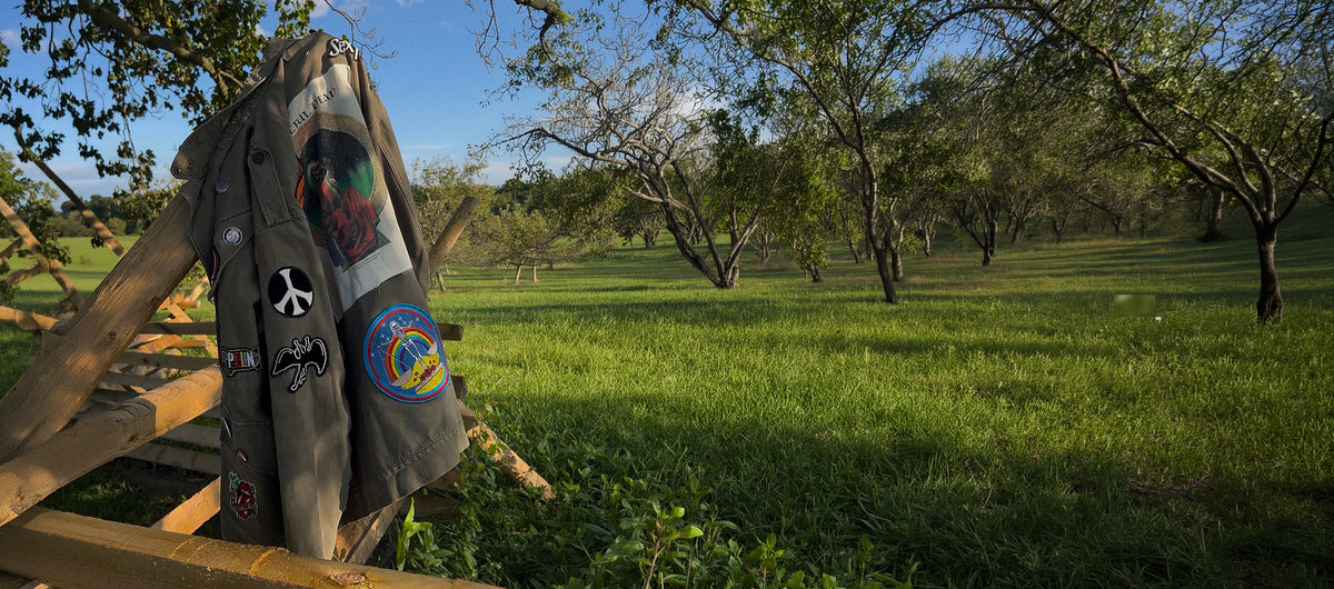 Patched military jacket hanging over a wooden fence in a green field.